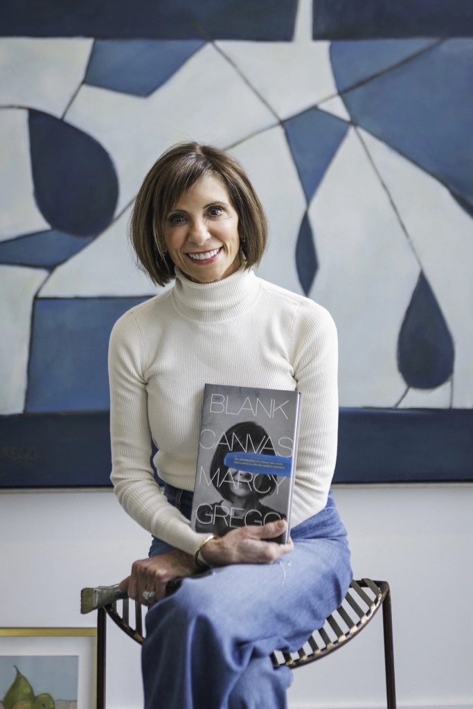 Author Marcy Gregg holding her memoir Blank Canvas, seated in front of one of her paintings